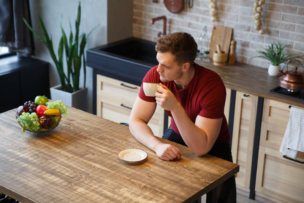 Gündelik giysiler içinde genç ve çekici bir adam mutfaktaki sıcak bir evde bir fincan kahve ile oturuyor.. - Fotoğraf, Görsel