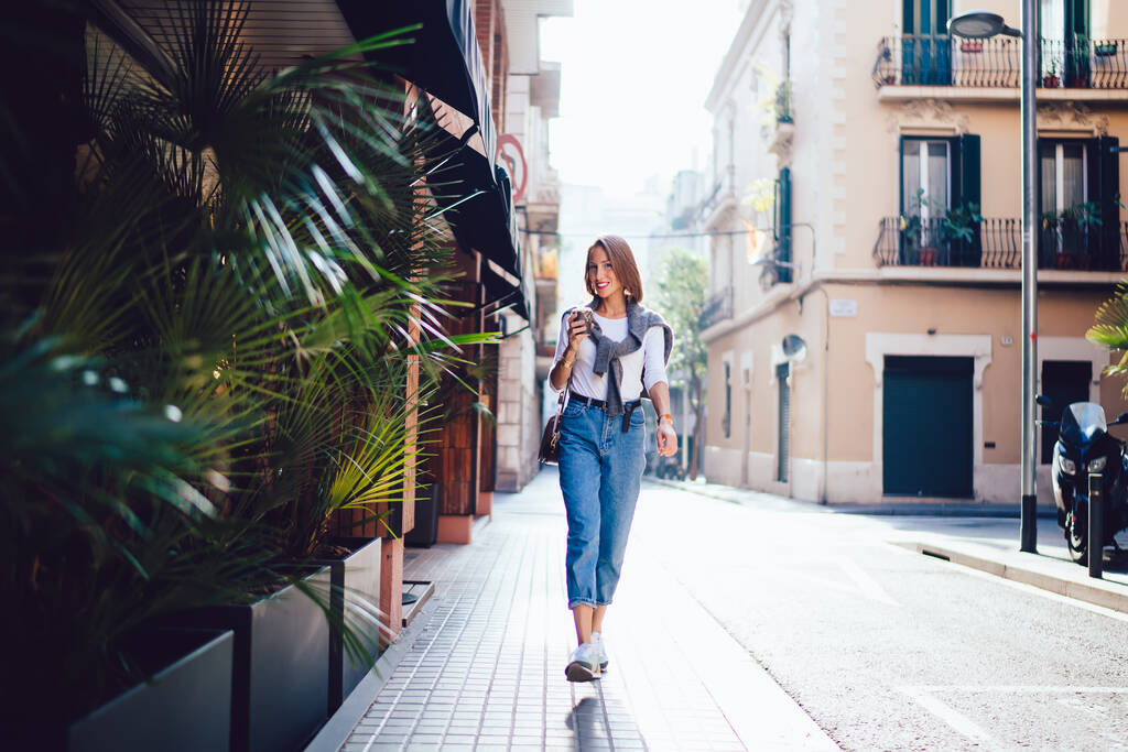 Corpo cheio de positivo sorridente feminino pedestre em roupa casual passeando na rua vazia perto de cafés modernos na manhã ensolarada - Foto, Imagem