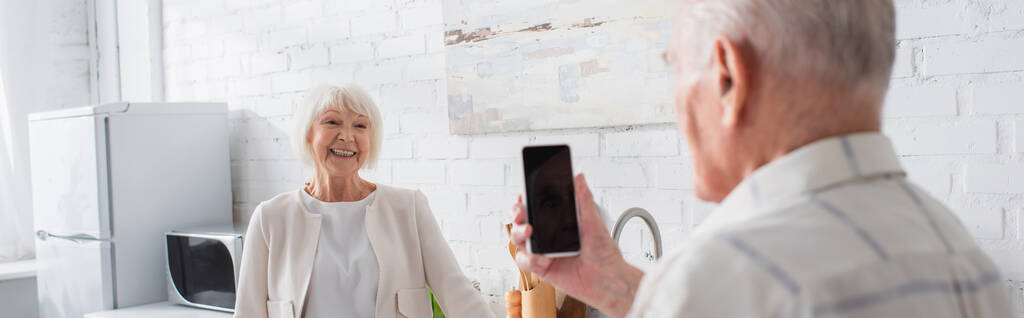 Hombre mayor sosteniendo teléfono inteligente con pantalla en blanco cerca de un amigo sonriente en el hogar de ancianos, pancarta  - Foto, imagen