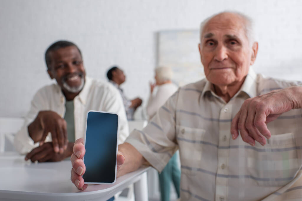 Smartphone na mão de homem sênior desfocado perto de amigo afro-americano em lar de idosos  - Foto, Imagem