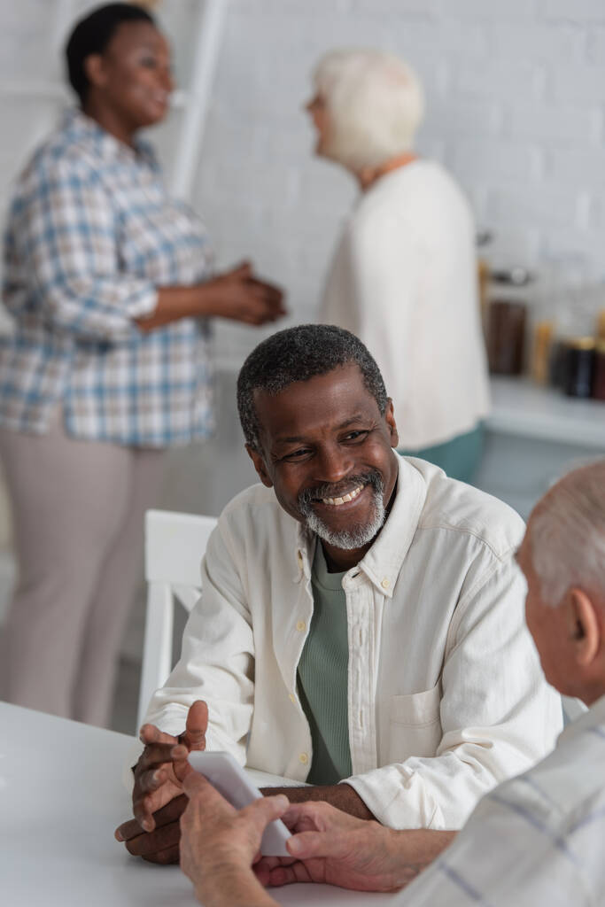 glimlachende Afrikaan amerikaanse man op zoek naar senior vriend met smartphone in verpleeghuis  - Foto, afbeelding