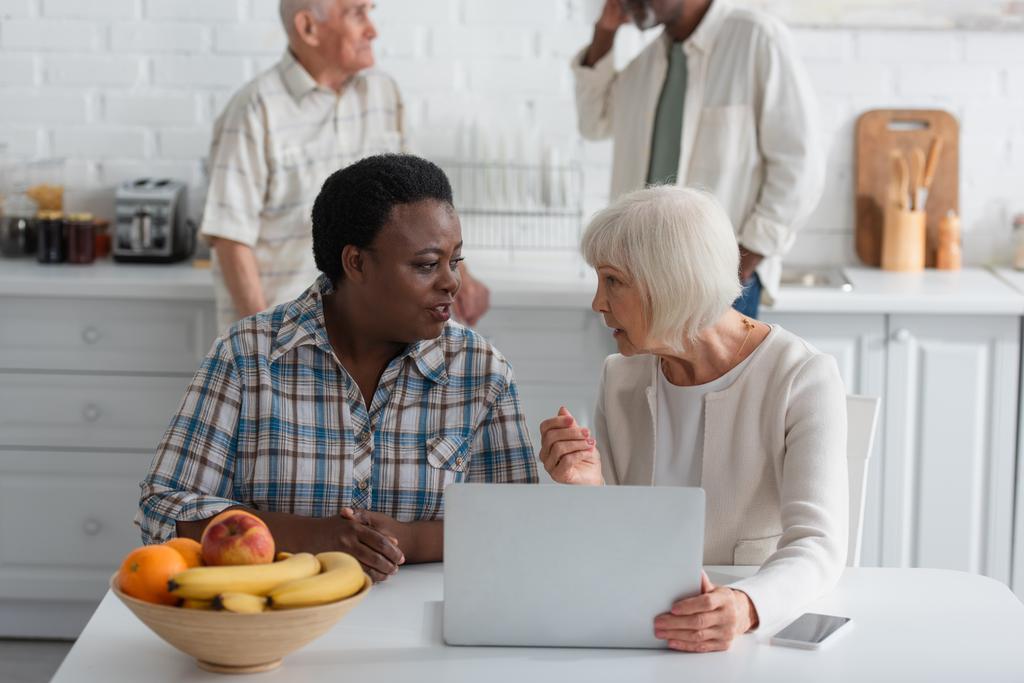 Senior interracial women talking near devices and fruits in nursing home  - Photo, Image