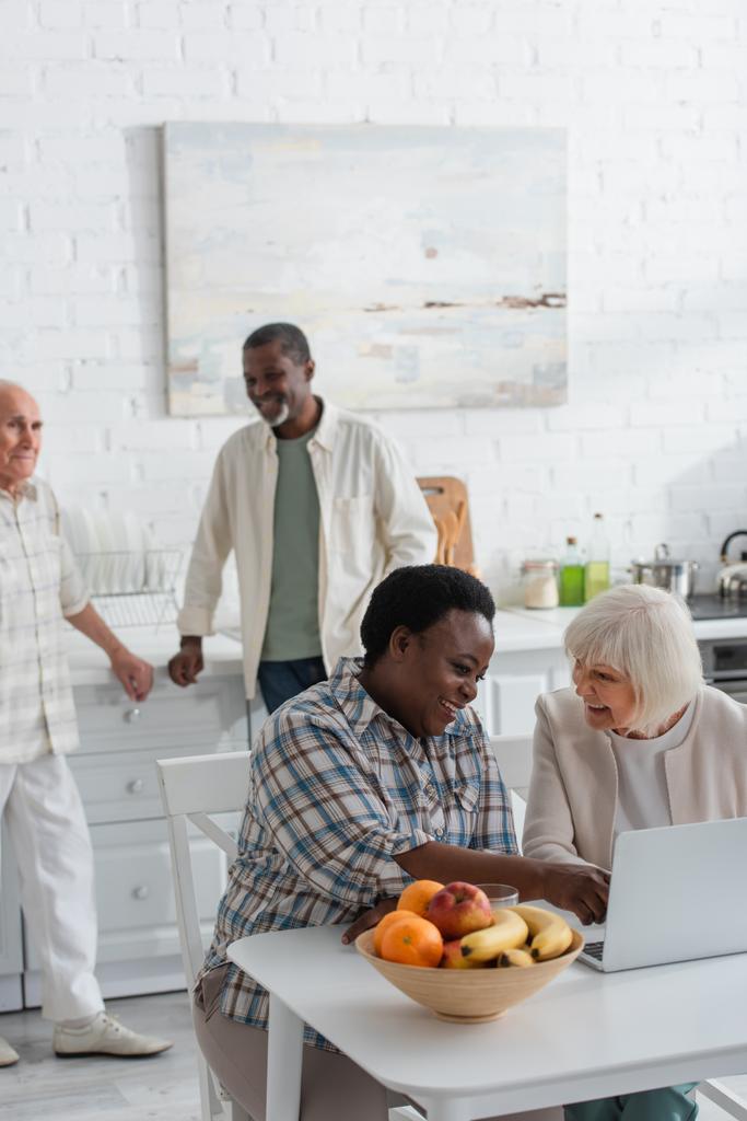 Mosolygó többnemzetiségű nők laptop közelében gyümölcsök idősek otthonában  - Fotó, kép