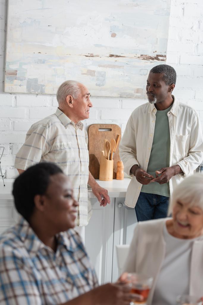 Ancianos hombres interracial hablando cerca de las mujeres borrosas en el hogar de ancianos  - Foto, Imagen