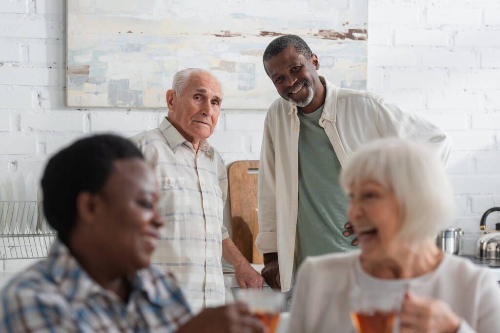 Hombres multiétnicos mayores mirando a la cámara cerca de mujeres sosteniendo té en un asilo de ancianos  - Foto, imagen