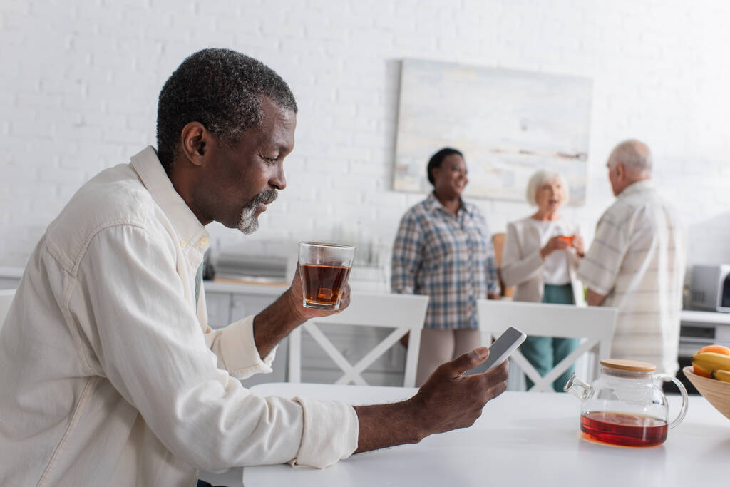 Uśmiechnięty starszy Afroamerykanin z herbatą przy użyciu smartfona w domu opieki  - Zdjęcie, obraz