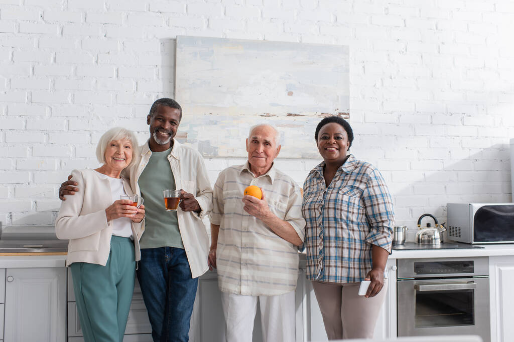 Χαρούμενη πολυεθνική ηλικιωμένους που κατέχουν τσάι και smartphone στην κουζίνα στο γηροκομείο  - Φωτογραφία, εικόνα