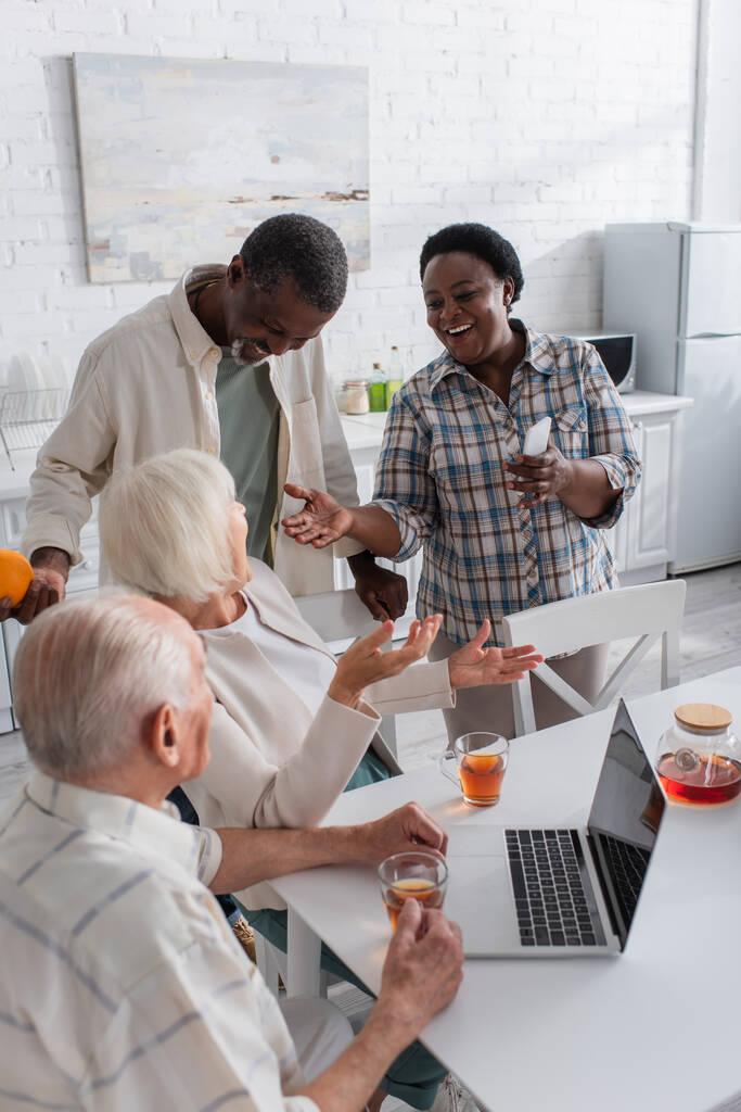 Glückliche Afroamerikanerin hält Smartphone in der Hand, während sie mit Seniorfreundin in der Nähe von Laptop und Tee im Pflegeheim spricht  - Foto, Bild