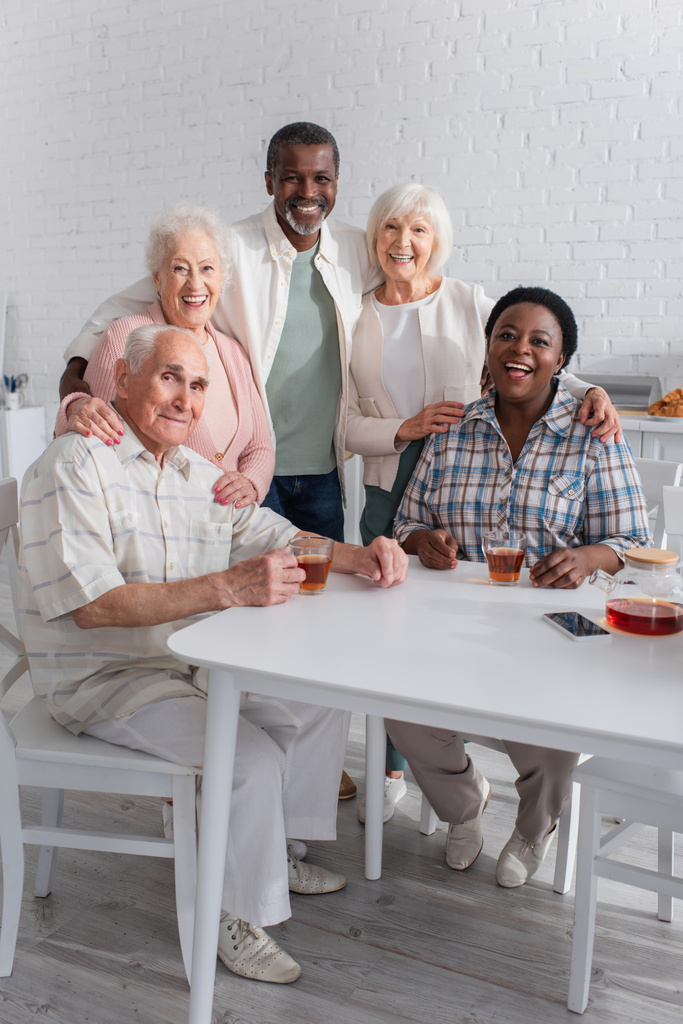 Pozitivní multietnické senioři objímání v blízkosti smartphone a čaj v pečovatelském domě  - Fotografie, Obrázek