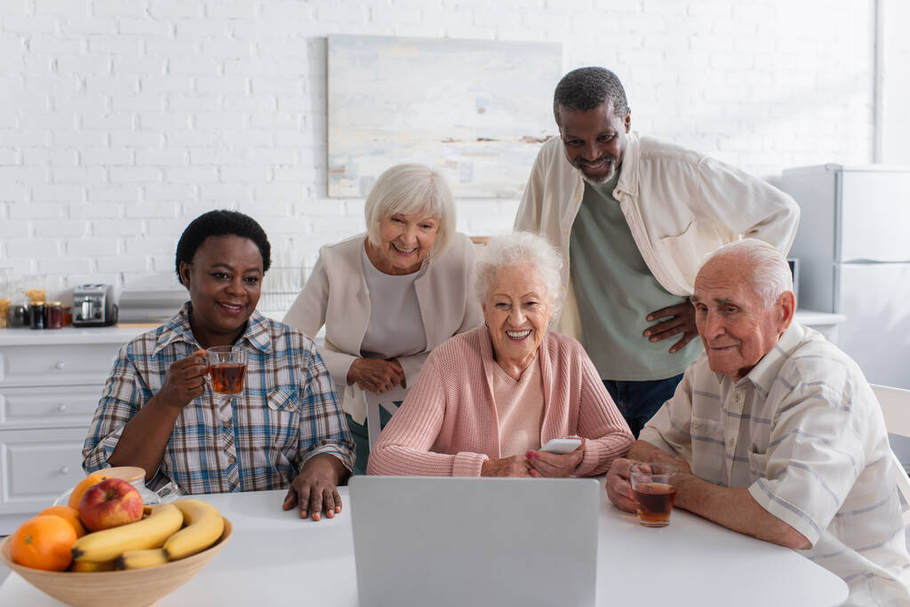 Χαρούμενοι πολυεθνικοί φίλοι που χρησιμοποιούν συσκευές κοντά στο τσάι στο γηροκομείο  - Φωτογραφία, εικόνα