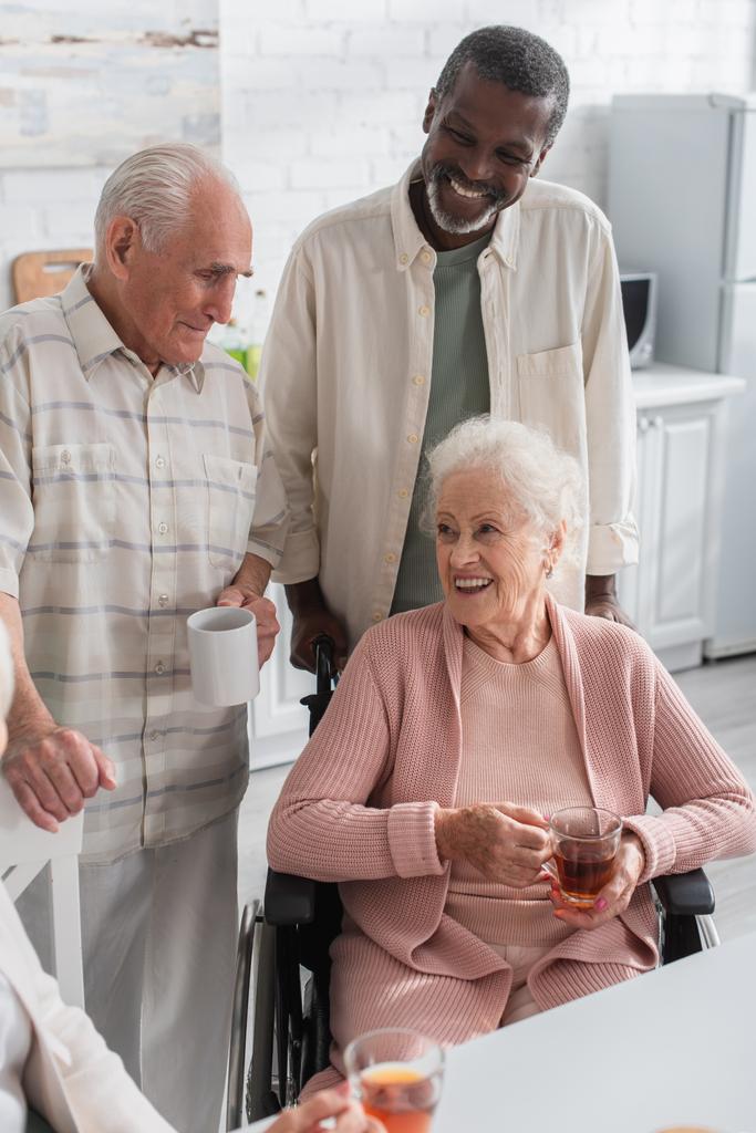 Elderly interracial men with tea standing near friend in wheelchair in nursing home  - Photo, Image