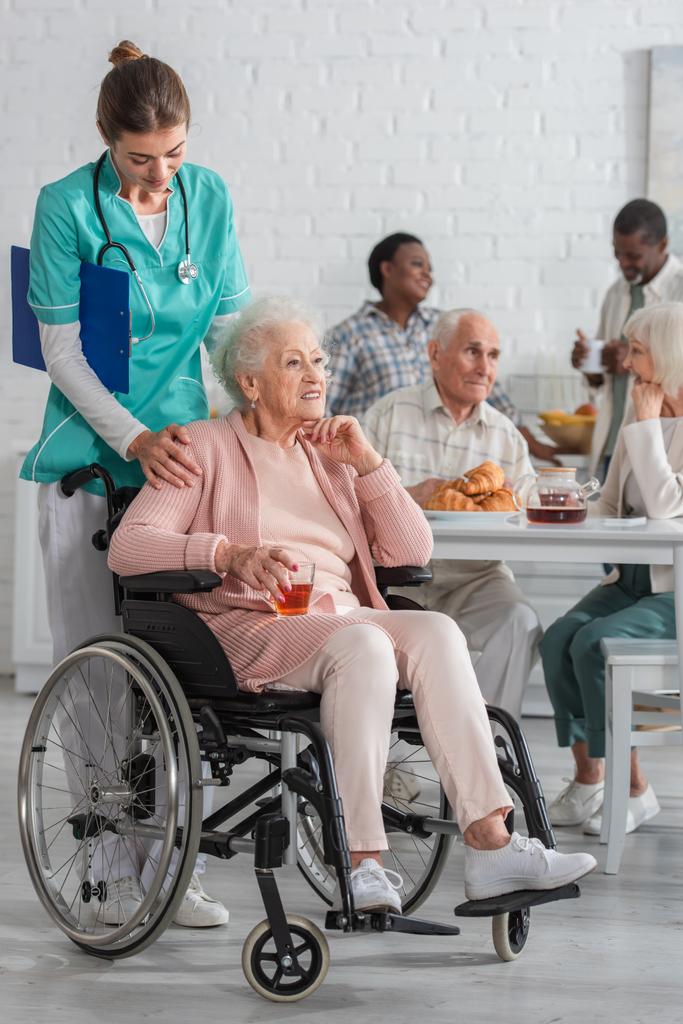 Bakımevinde tekerlekli sandalyede çay içen bir hastanın yanında duran hasta bakıcı.  - Fotoğraf, Görsel