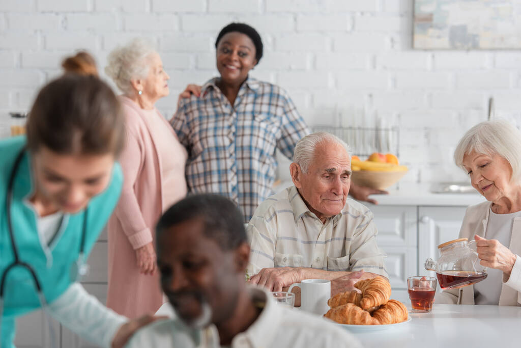 Senior nainen tilalla teekannu lähellä ystävä ja hämärtynyt rotujenvälinen ihmisiä hoitokodissa  - Valokuva, kuva