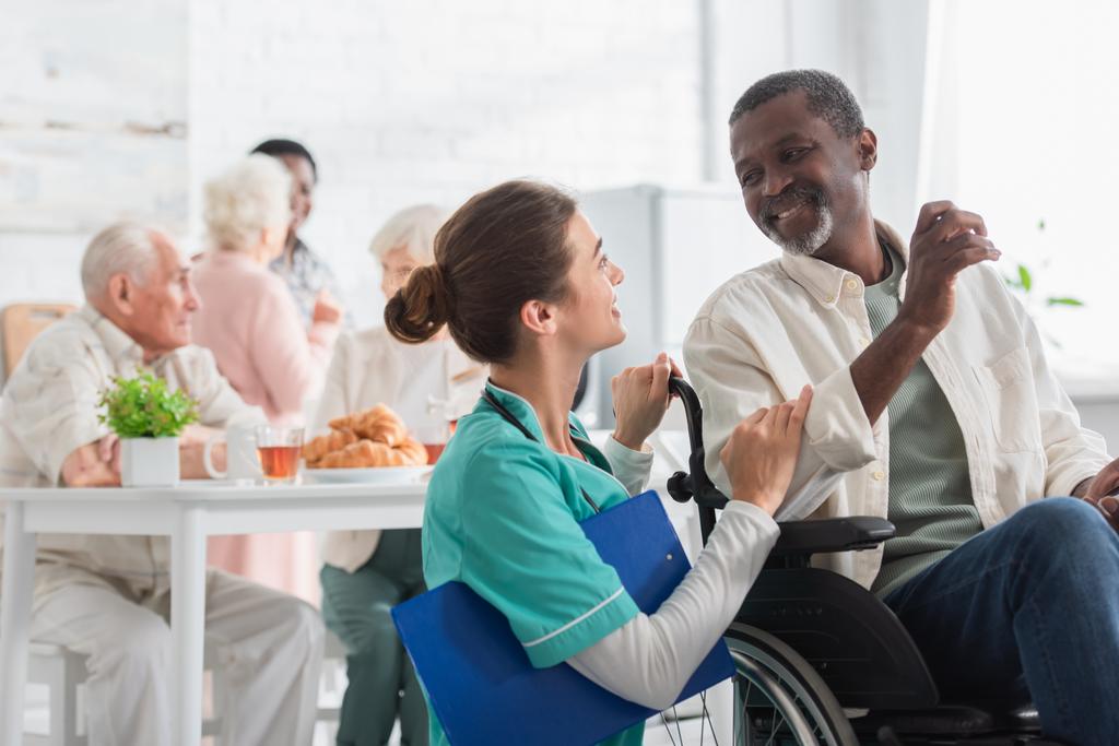 Vrolijke verpleegster met klembord praten met Afro-Amerikaanse man in rolstoel in verpleeghuis  - Foto, afbeelding