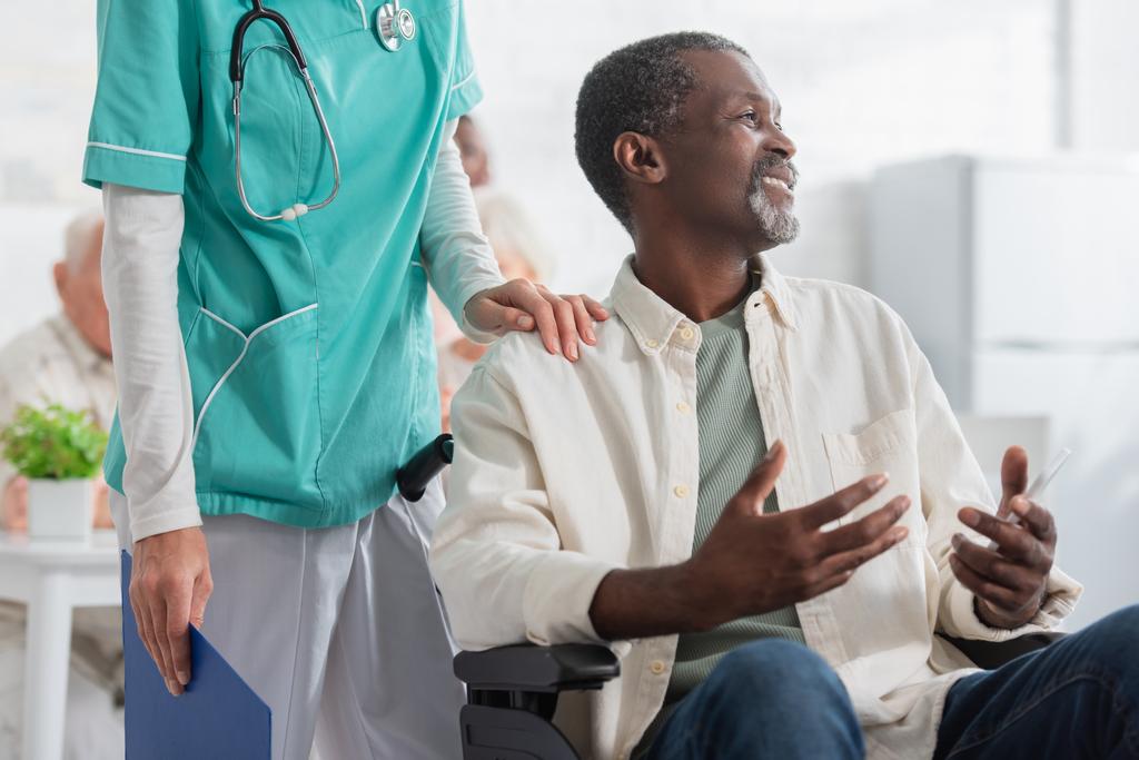 Усміхнений афроамериканський пацієнт тримає смартфон сидячи в інвалідному візку біля медсестри в будинку престарілих
  - Фото, зображення