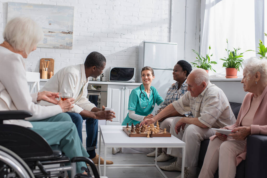 Infermiera sorridente trascorrere del tempo con persone multietniche giocare a scacchi in casa di cura  - Foto, immagini