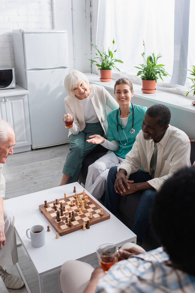 笑顔看護師ポインティングとともに手近く異人種間の患者とチェスで老人ホーム  - 写真・画像