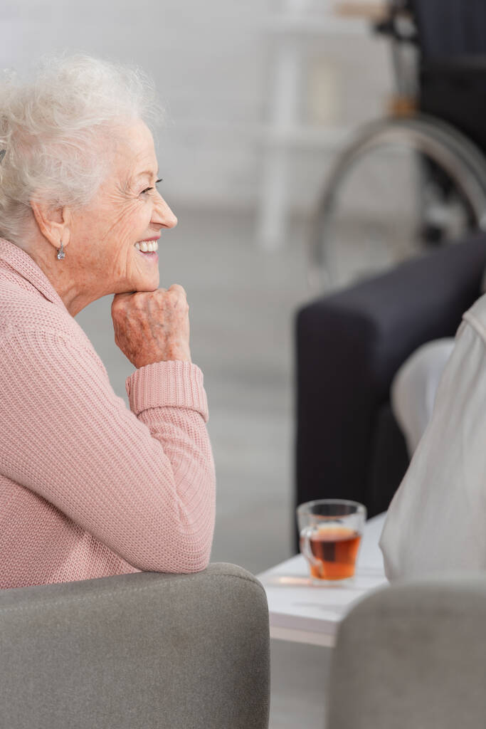 Seitenansicht einer lächelnden Seniorin, die neben verschwommenem Tee im Pflegeheim sitzt  - Foto, Bild