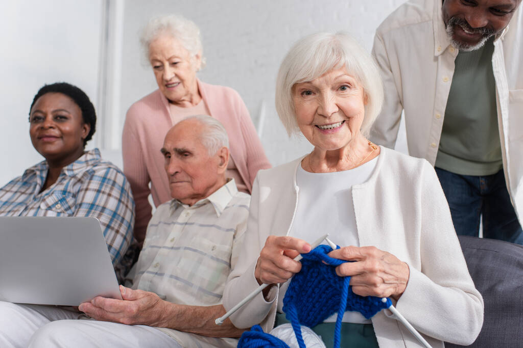 Mujer anciana sosteniendo hilo cerca de amigos multiétnicos con portátil en el hogar de ancianos  - Foto, Imagen
