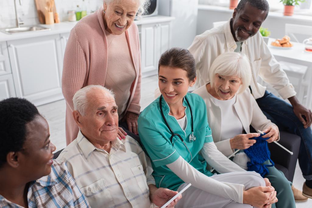 Hombre mayor sosteniendo tableta digital cerca de la enfermera y amigos interracial en el hogar de ancianos  - Foto, Imagen