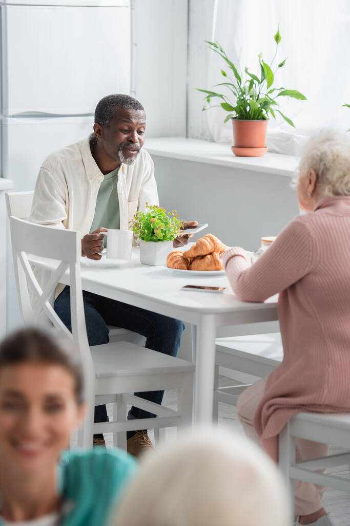 Homme afro-américain avec thé et smartphone parlant à un ami dans une maison de retraite  - Photo, image