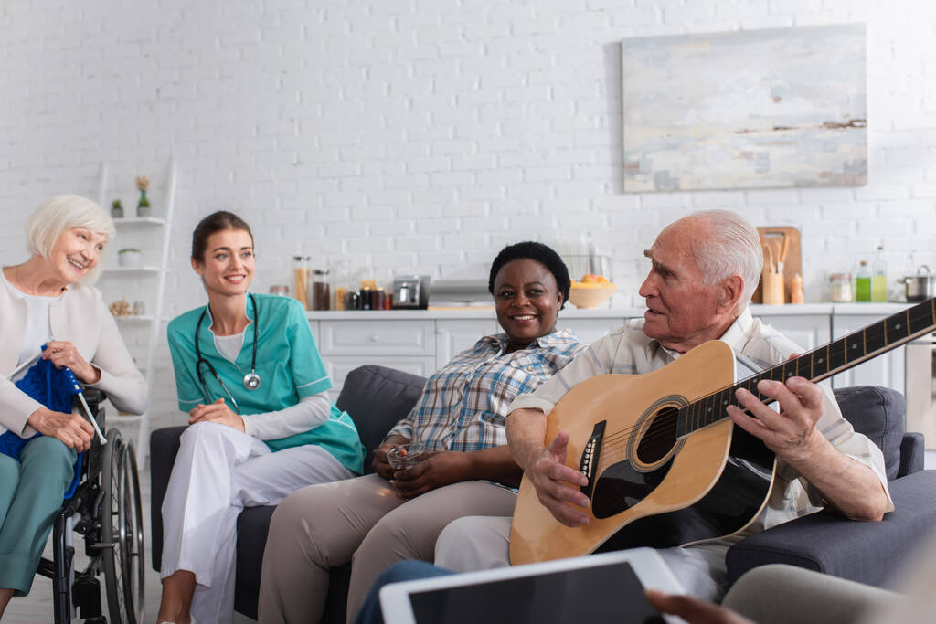 Idős férfi kezében akusztikus gitár közelében multietnikai barátok és a nővér az idősek otthonában  - Fotó, kép