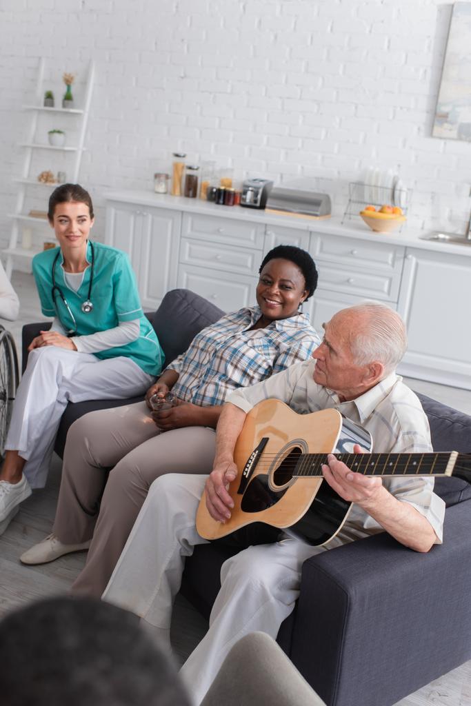 Пожилой человек играет на акустической гитаре рядом с африканским американским другом с чаем и медсестрой в доме престарелых  - Фото, изображение