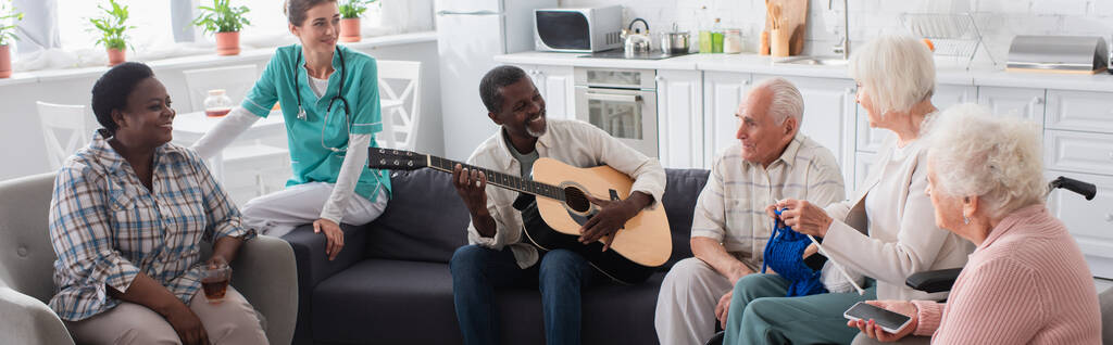 Hymyilevä rotujenvälinen vanhukset soittavat akustista kitaraa lähellä sairaanhoitaja hoitokodissa, banneri  - Valokuva, kuva