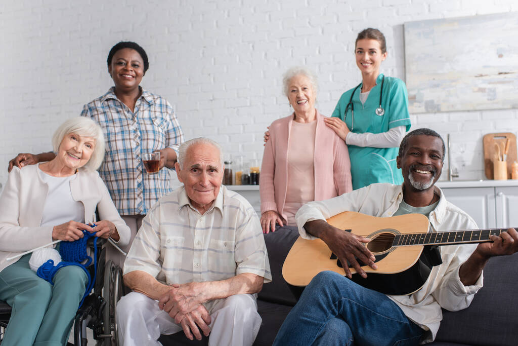 Θετικοί διαφυλετικοί συνταξιούχοι με νήματα και ακουστική κιθάρα κοιτάζοντας την κάμερα κοντά σε νοσηλευτές στο γηροκομείο  - Φωτογραφία, εικόνα
