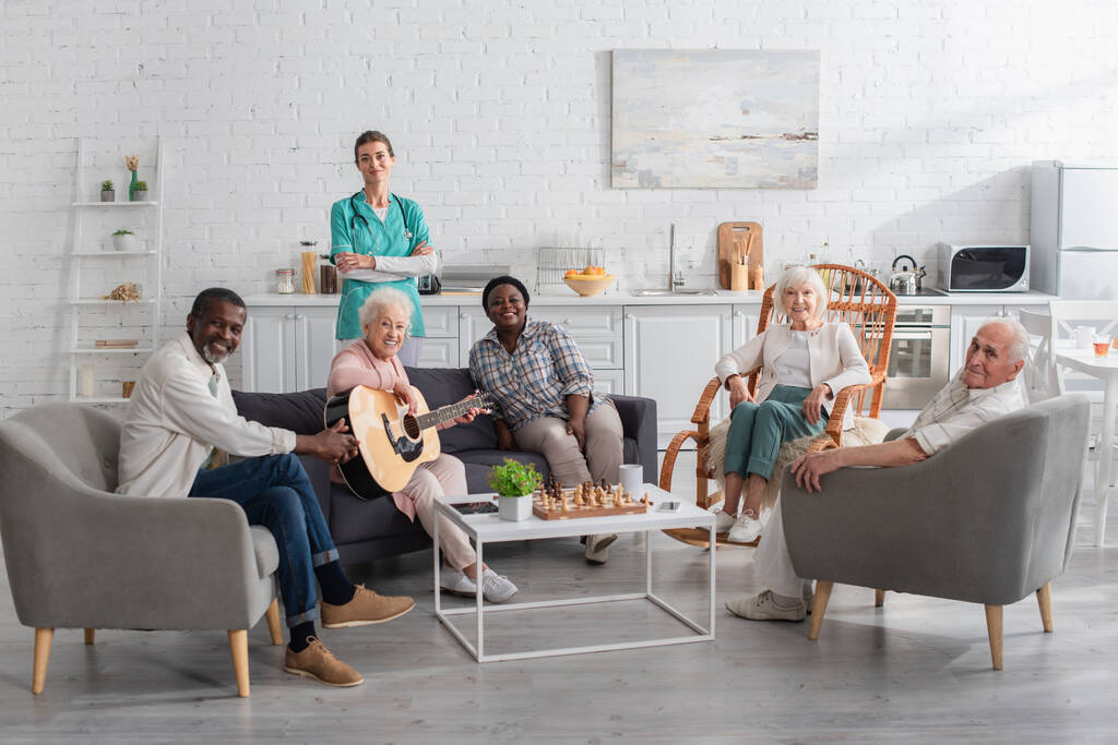 Vrolijke interraciale mensen met akoestische gitaar kijken naar camera in de buurt van schaken en verpleegster in verpleeghuis  - Foto, afbeelding