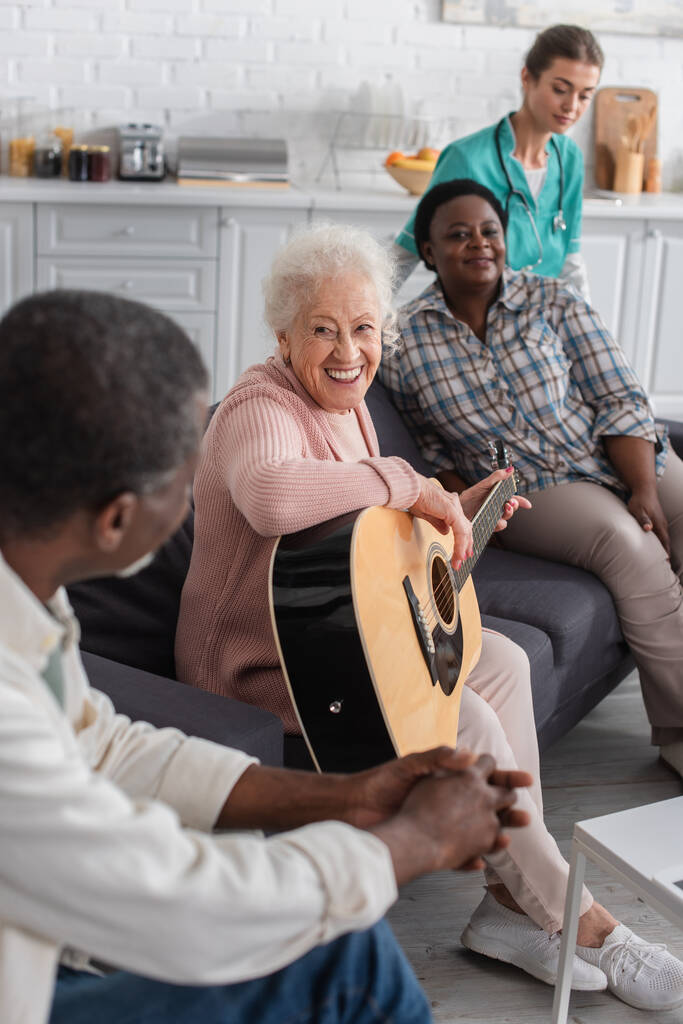 Улыбающаяся пожилая женщина, играющая на акустической гитаре рядом с многонациональными друзьями и размытая медсестра в доме престарелых  - Фото, изображение