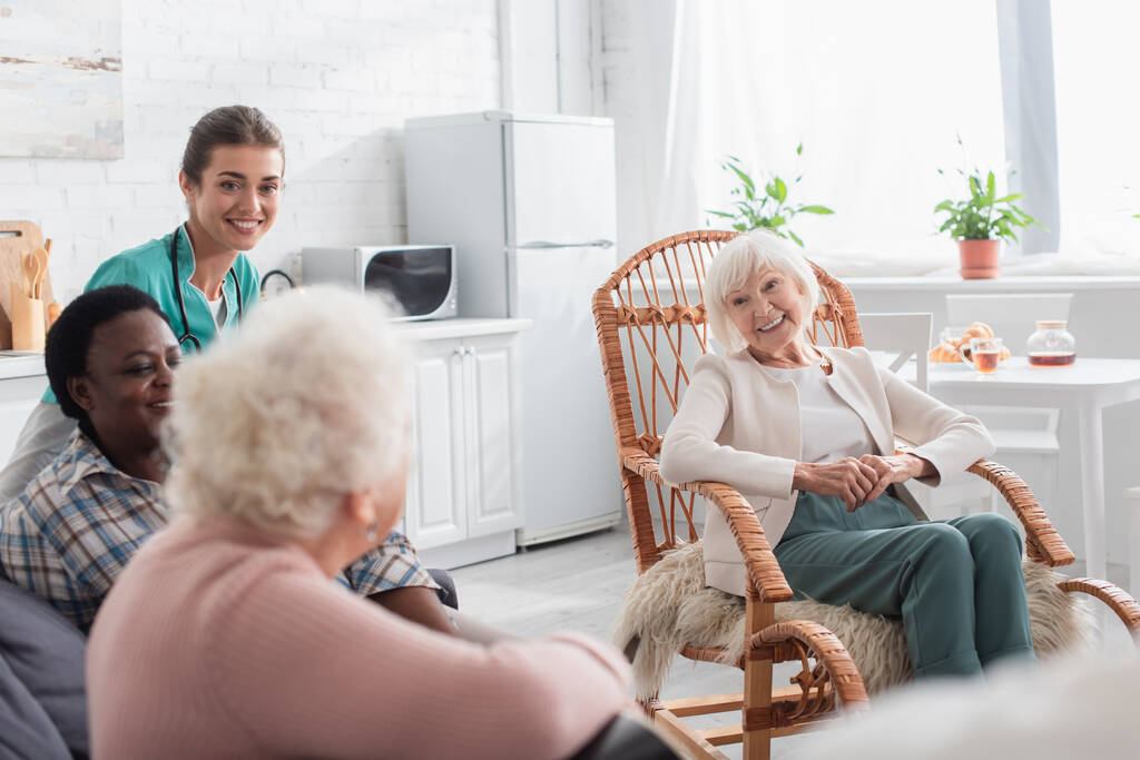 Cheerful senior interracial women spending time near young nurse in nursing home  - Photo, Image