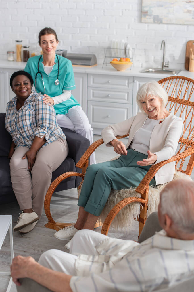 Багатоетнічні літні люди розмовляють біля медсестри на дивані в будинку престарілих
  - Фото, зображення