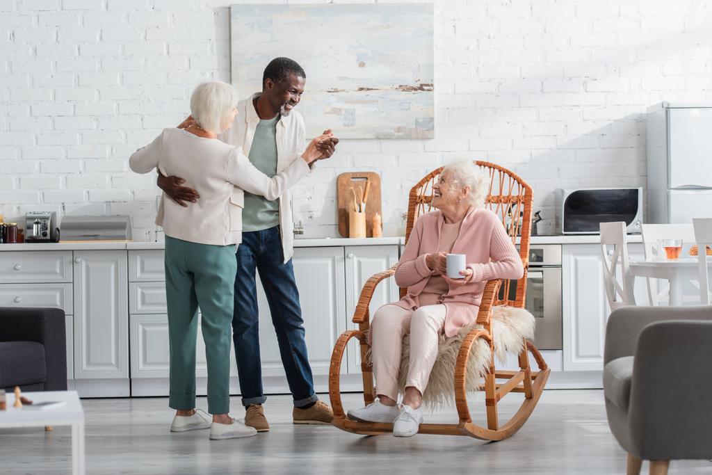 Mujer mayor sonriente sosteniendo taza en mecedora cerca de amigos interracial bailando en un hogar de ancianos  - Foto, imagen