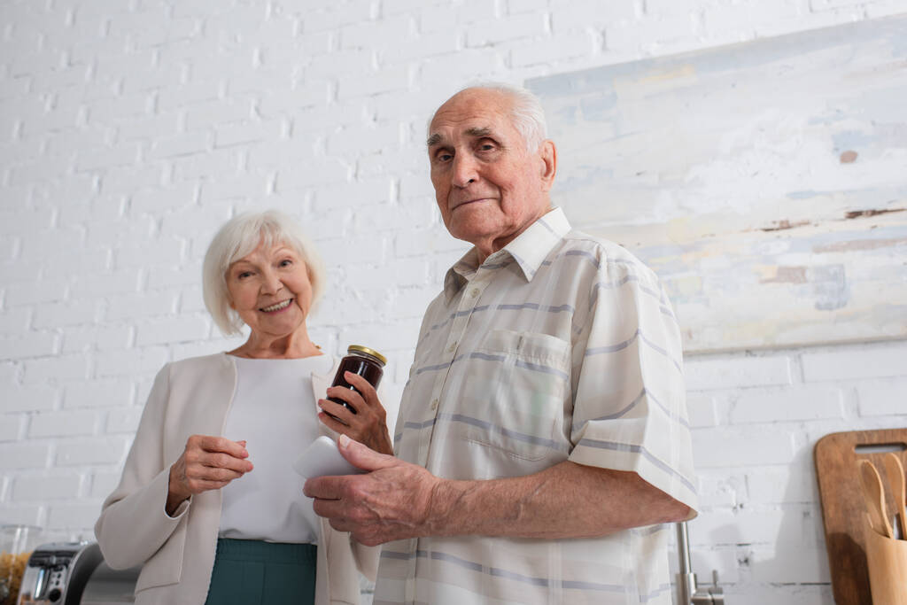 Χαμηλή γωνία άποψη του ηλικιωμένου άνδρα κρατώντας smartphone κοντά σε φίλο με βάζο μαρμελάδα στο γηροκομείο  - Φωτογραφία, εικόνα