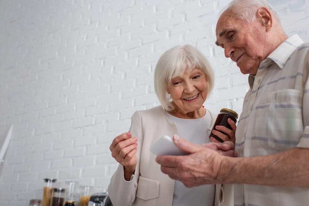 Starszy mężczyzna pokazuje smartfon uśmiechniętemu przyjacielowi ze słoikiem dżemu w domu opieki  - Zdjęcie, obraz