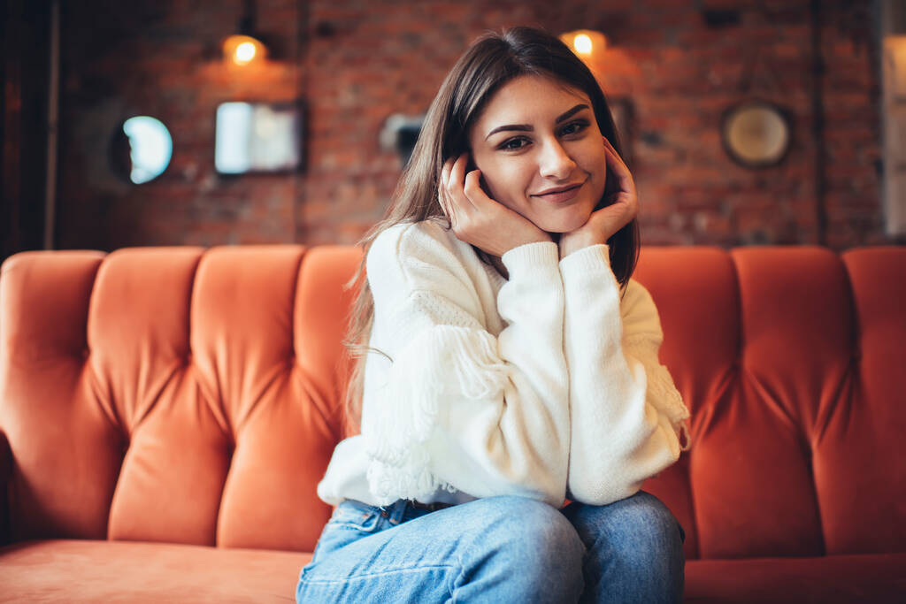Satisfied female in casual sweater and jeans looking at camera while sitting on comfortable couch in light cafe leaning on hands - Photo, Image
