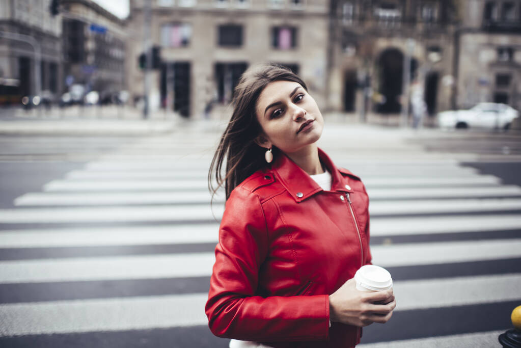 Joven mujer reflexiva en ropa informal brillante con el pelo largo mirando a la cámara mientras está de pie cerca del paso de peatones con taza de papel de café - Foto, imagen