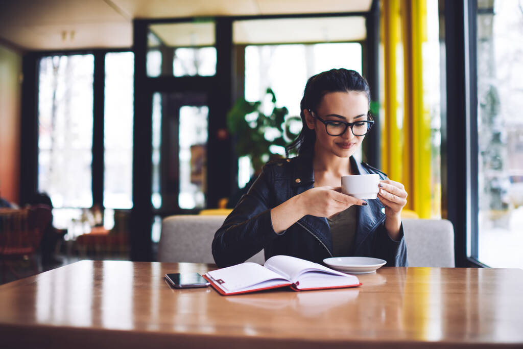 Ηρεμία γυναίκα ελεύθερος επαγγελματίας κάθεται στο τραπέζι στο φως καφέ με ανοιχτή planner και απολαμβάνοντας καφέ, ενώ έχοντας διάλειμμα κατά τη διάρκεια της εργάσιμης ημέρας - Φωτογραφία, εικόνα