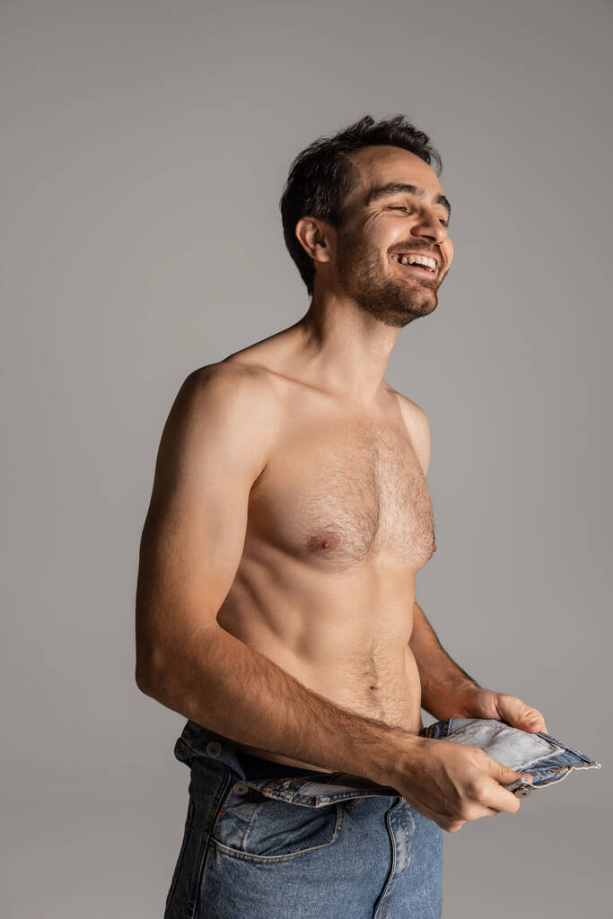 Retrato de jovem bonito barbudo sem camisa homem em jeans posando isolado no fundo do estúdio cinza. - Foto, Imagem