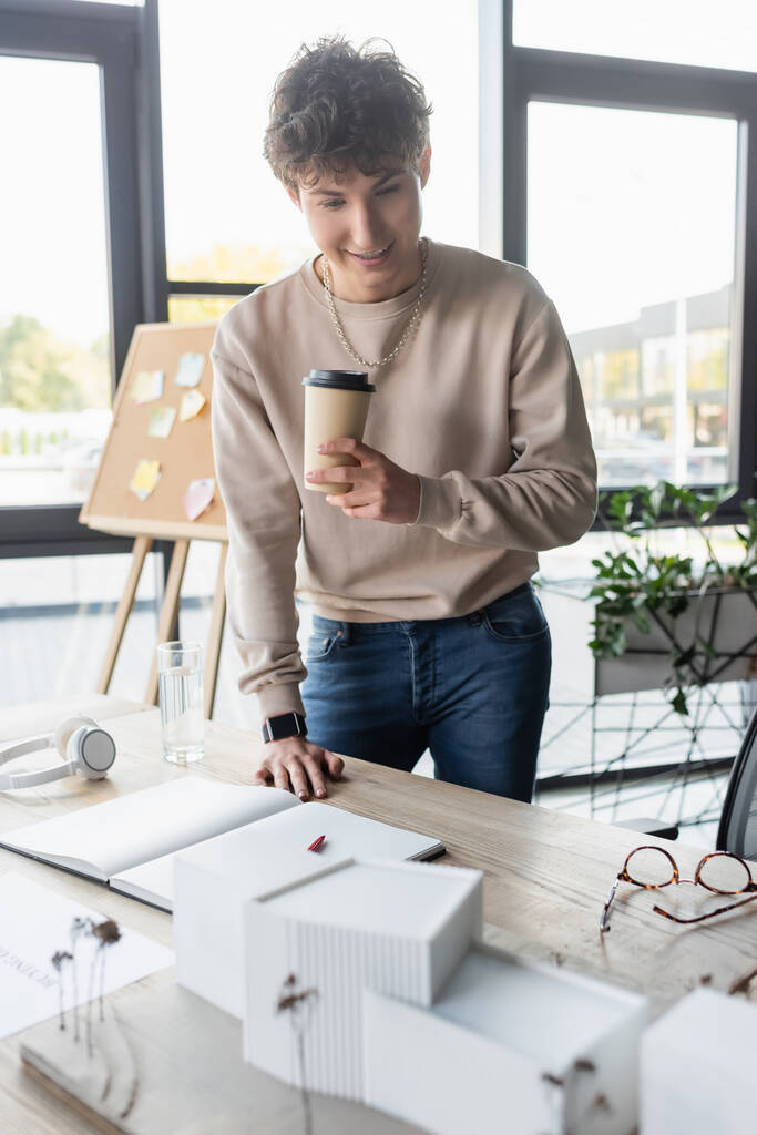 Pozitív üzletember kezében papír pohár és nézi modell épület közelében notebook az irodában  - Fotó, kép
