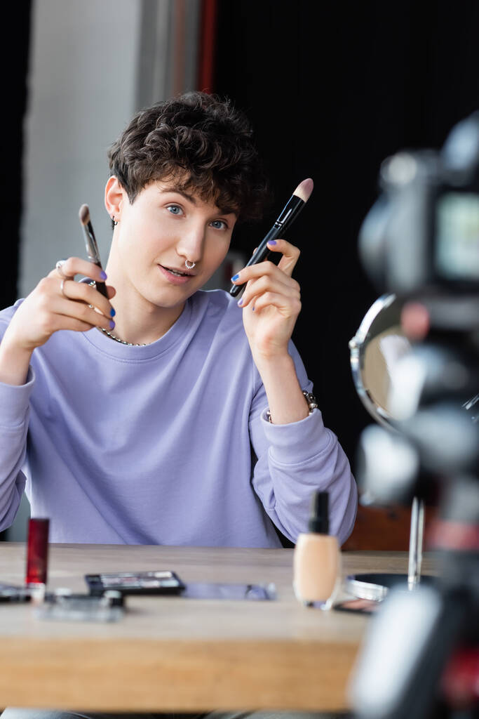 Transgender Make-up Artist hält Kosmetikpinsel in der Nähe verschwommener Digitalkamera  - Foto, Bild