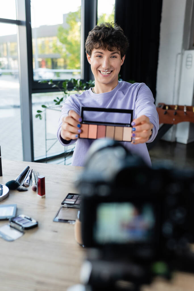 Sonriente artista de maquillaje transexual mostrando sombras de ojos en la cámara digital  - Foto, Imagen