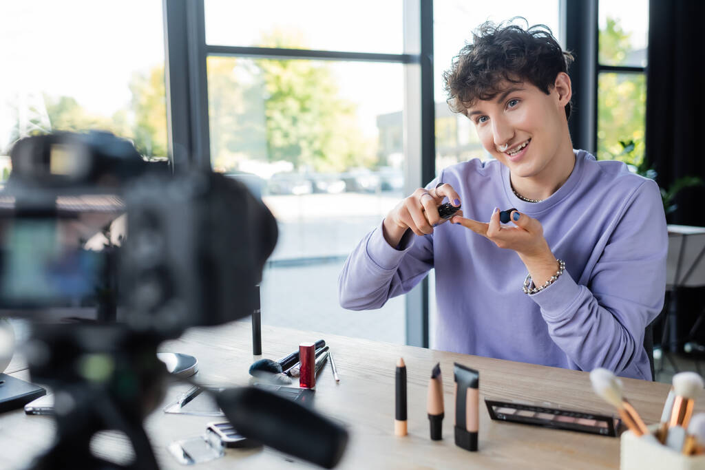 Gülümseyen transseksüel kişi dijital kameranın yanında kozmetik ürünler tutuyor.  - Fotoğraf, Görsel