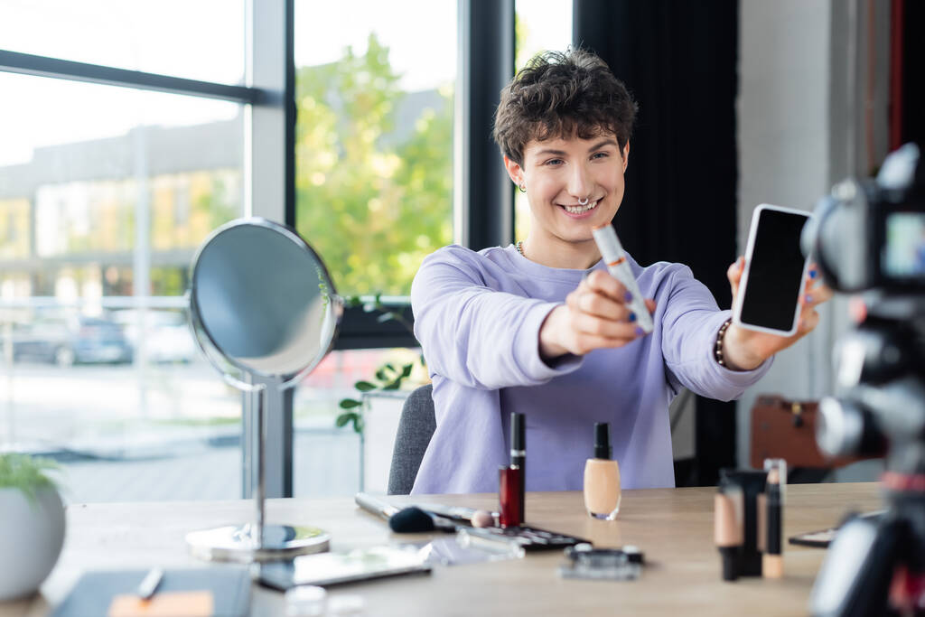 Uśmiechnięta transseksualistka trzymająca tusz do rzęs i smartfon w pobliżu kosmetyków dekoracyjnych i aparatu cyfrowego  - Zdjęcie, obraz