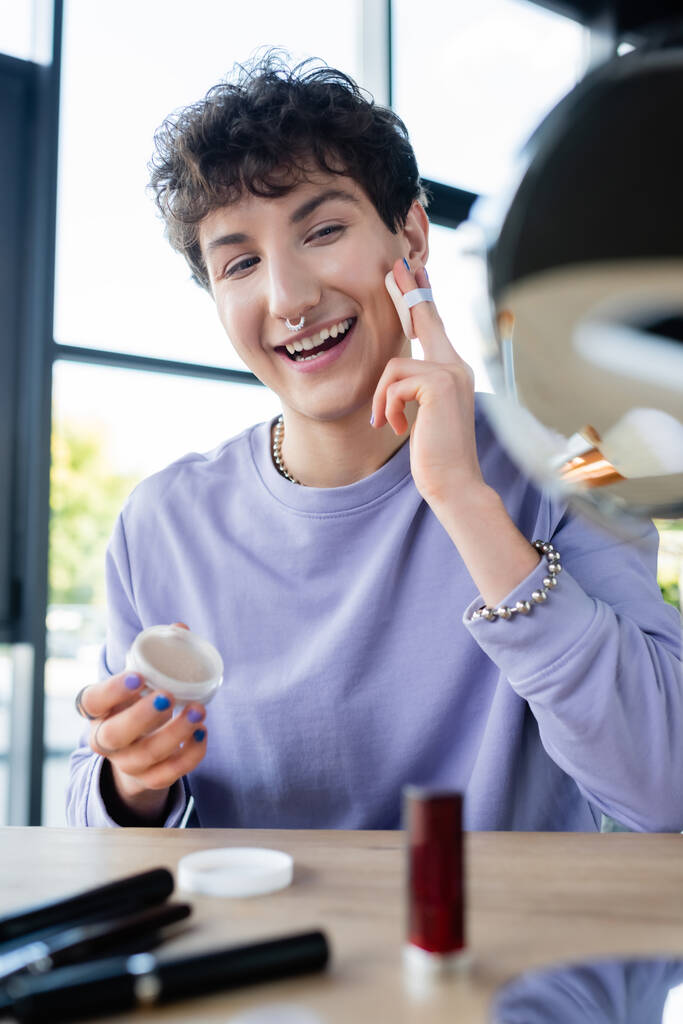 Persona transgénero feliz aplicando polvo facial cerca de cosméticos decorativos borrosos y espejo  - Foto, imagen