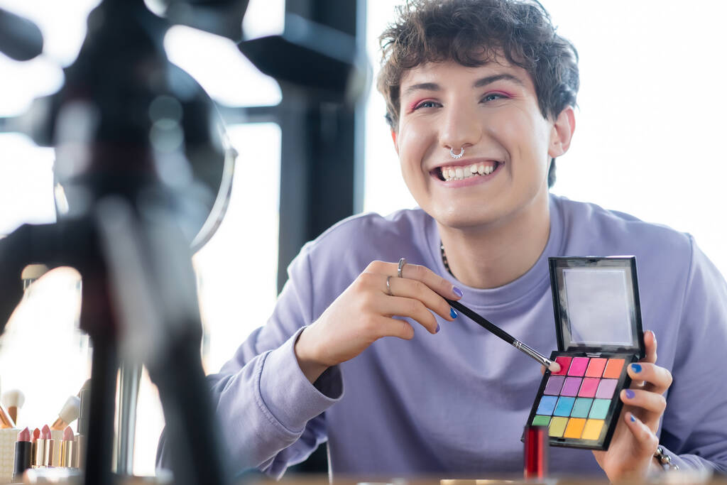 Hymyilevä transsukupuolinen henkilö, jolla on luomivärejä ja kosmeettinen harja kolmijalan ja peilin lähellä  - Valokuva, kuva