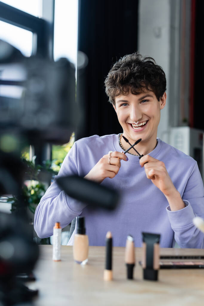 Sorrindo transgênero pessoa segurando escovas de rímel perto de cosméticos decorativos e câmera digital  - Foto, Imagem