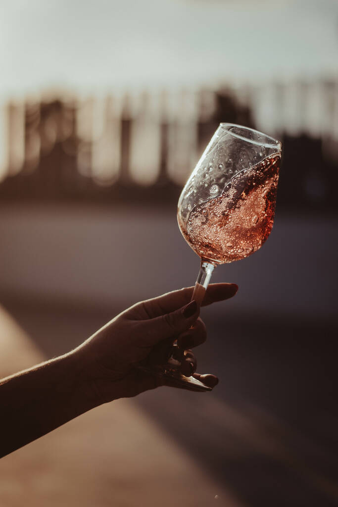 Вертикальний знімок скляного поля з трояндовим вином, що тримається жіночою рукою на розмитому фоні
 - Фото, зображення