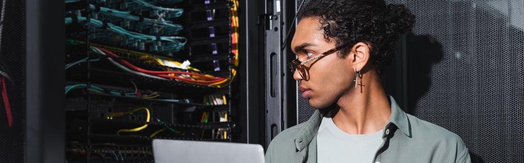 Afrykański inżynier amerykański z laptopem patrząc na przewody w serwerze podczas pracy w centrum danych, baner - Zdjęcie, obraz